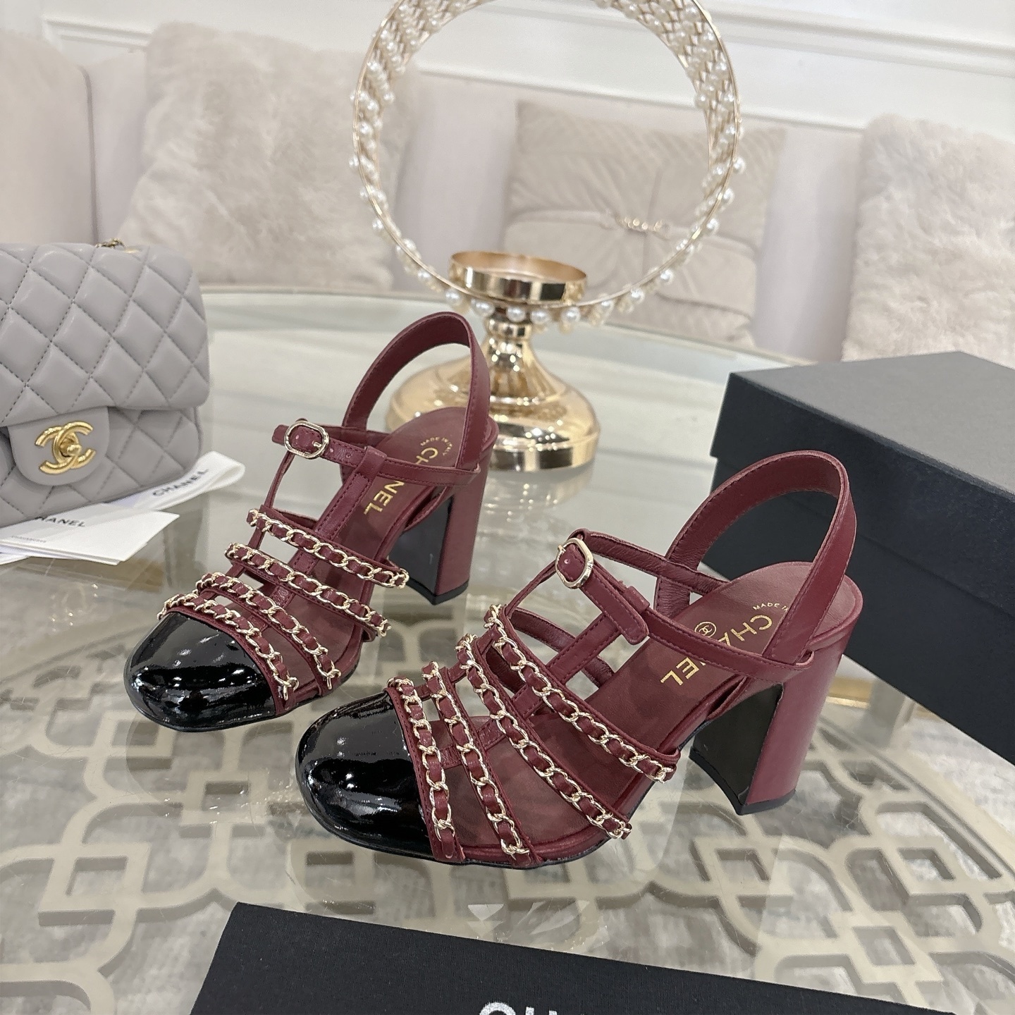 A pair of Chanel burgundy leather block heel sandals with gold chain straps and black patent cap toe displayed on a glass table. - Hình 3