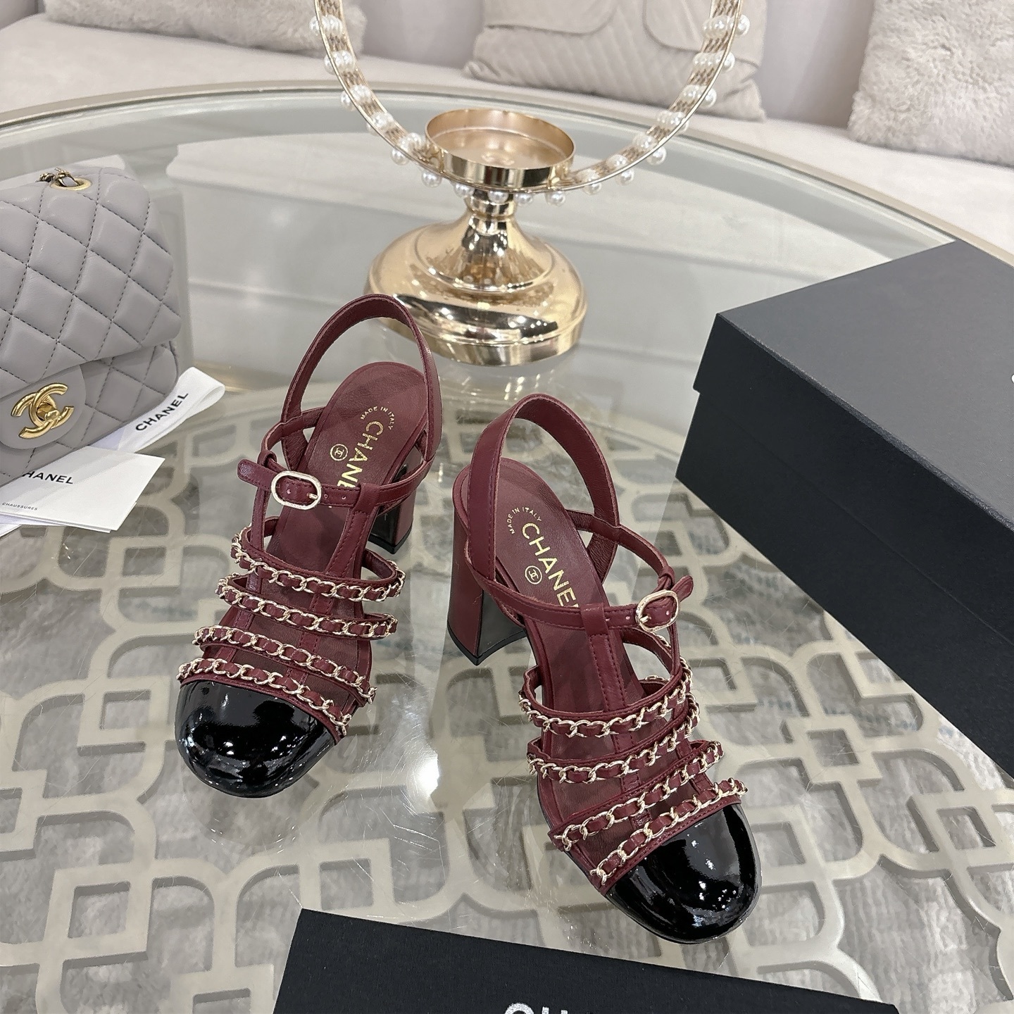 A pair of Chanel burgundy leather block heel sandals with gold chain straps and black patent cap toe displayed on a glass table. - Hình 2