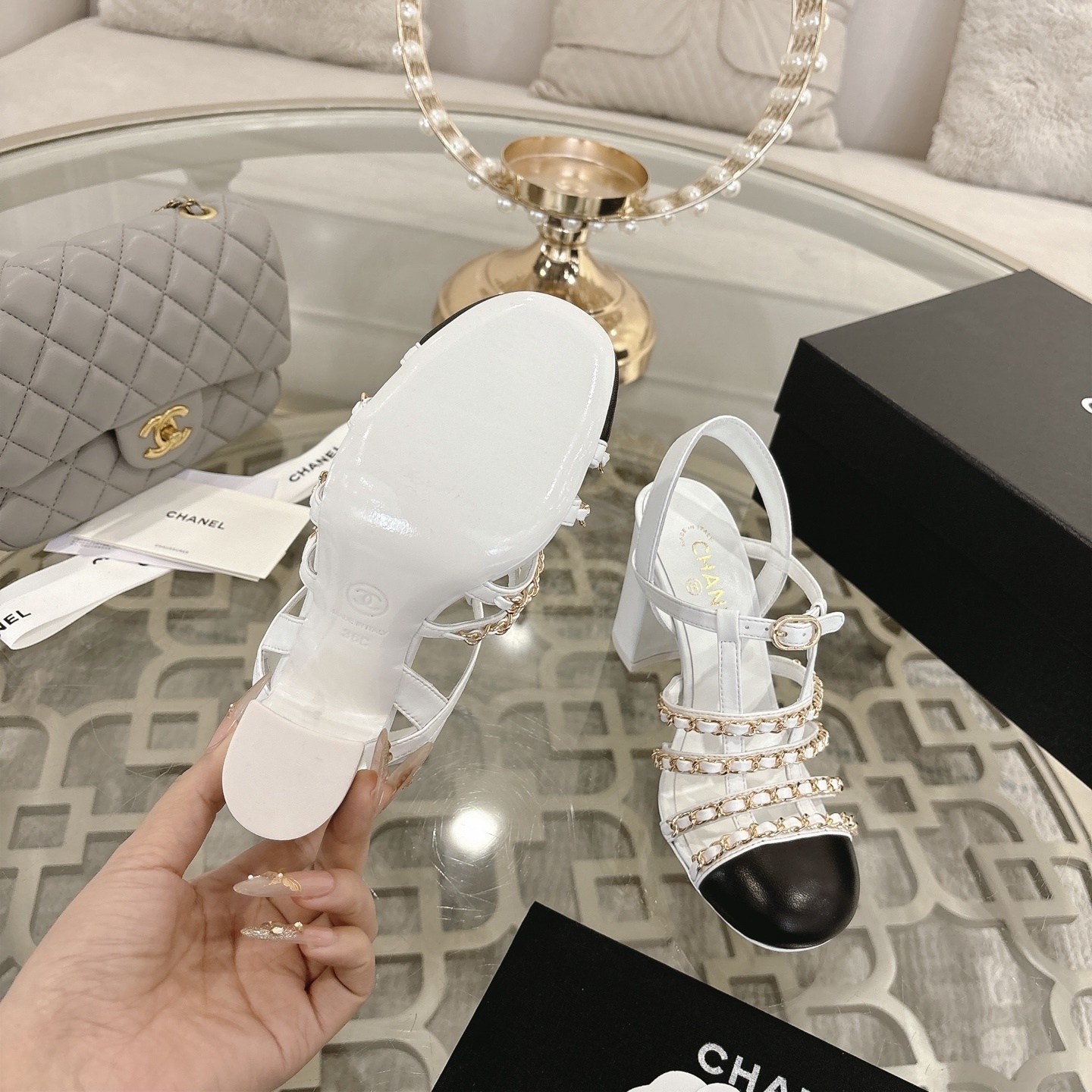Elegant white leather sandals with black cap-toes and gold chain straps displayed on a glass table. - Hình 9