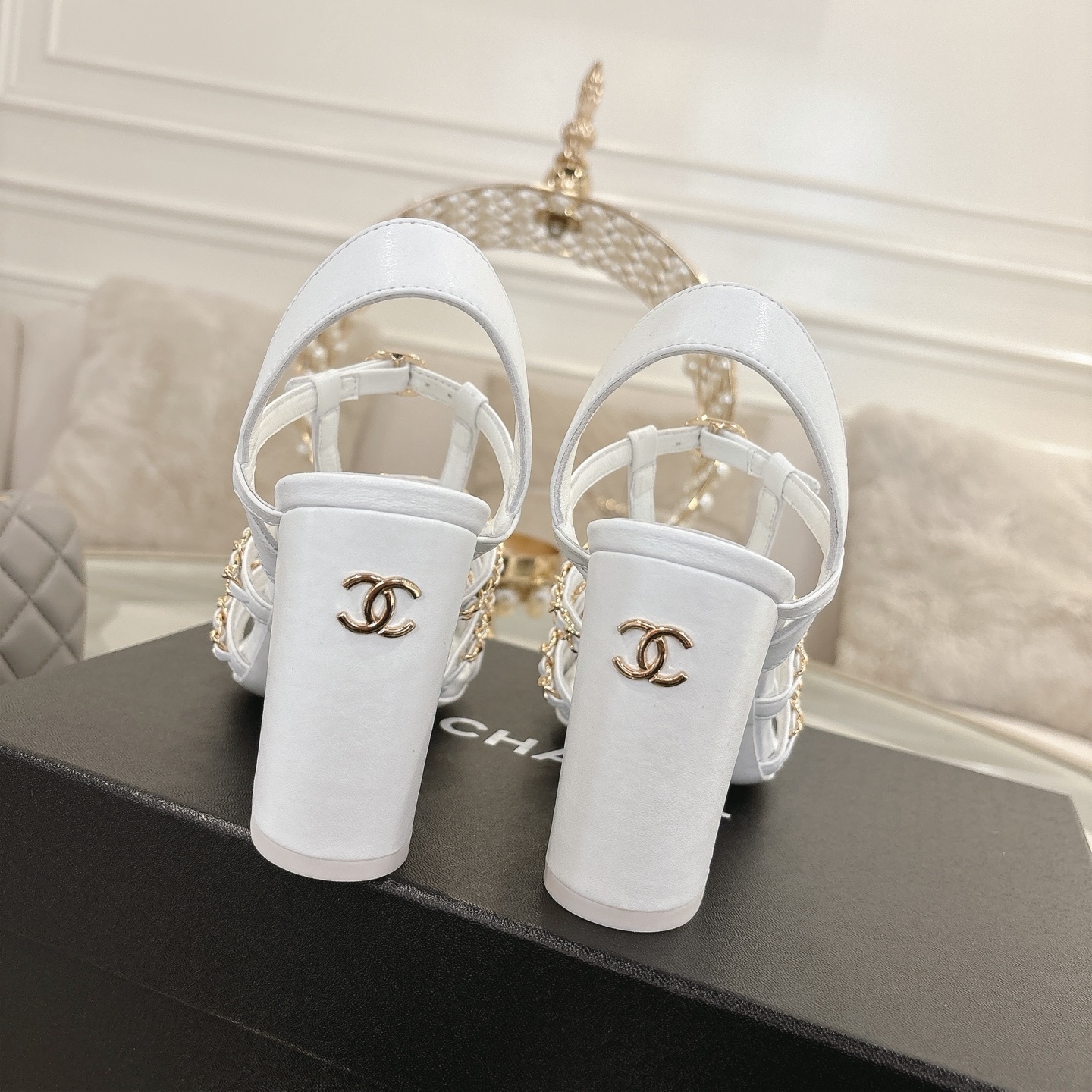 Elegant white leather sandals with black cap-toes and gold chain straps displayed on a glass table. - Hình 8