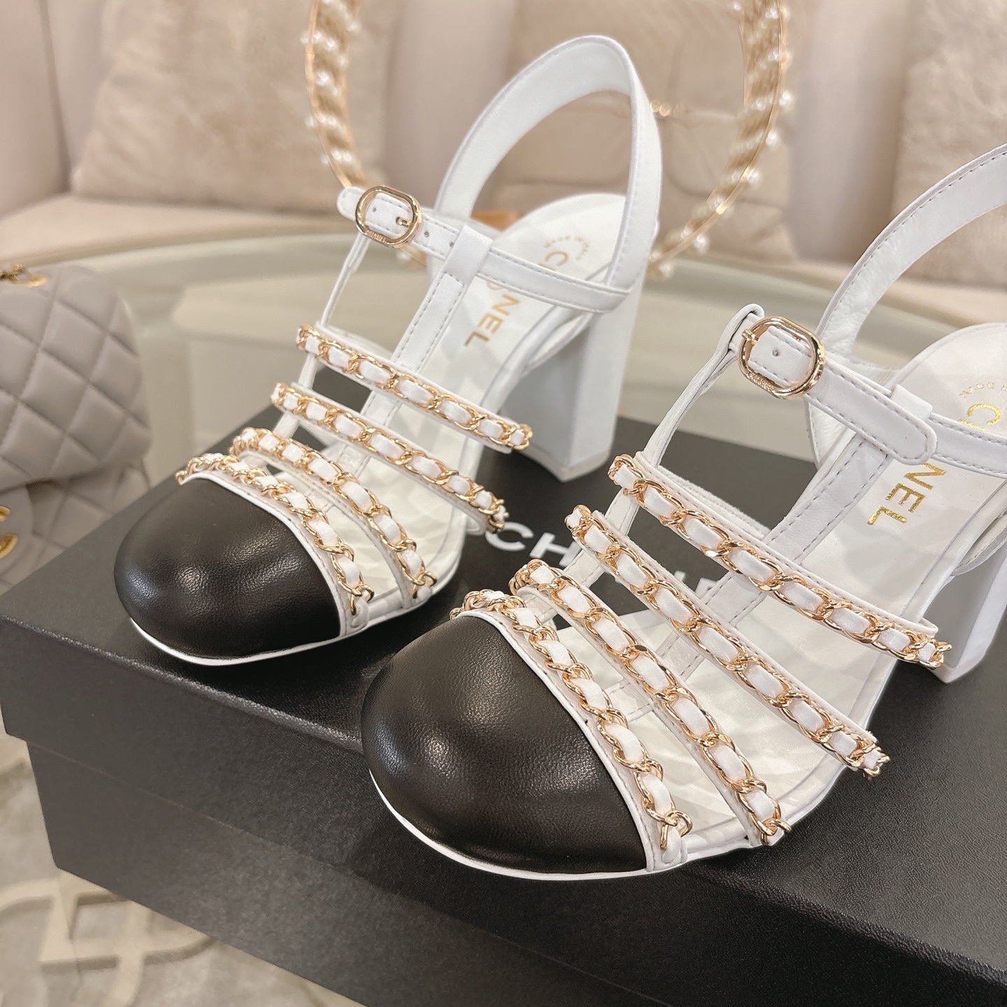 Elegant white leather sandals with black cap-toes and gold chain straps displayed on a glass table. - Hình 7