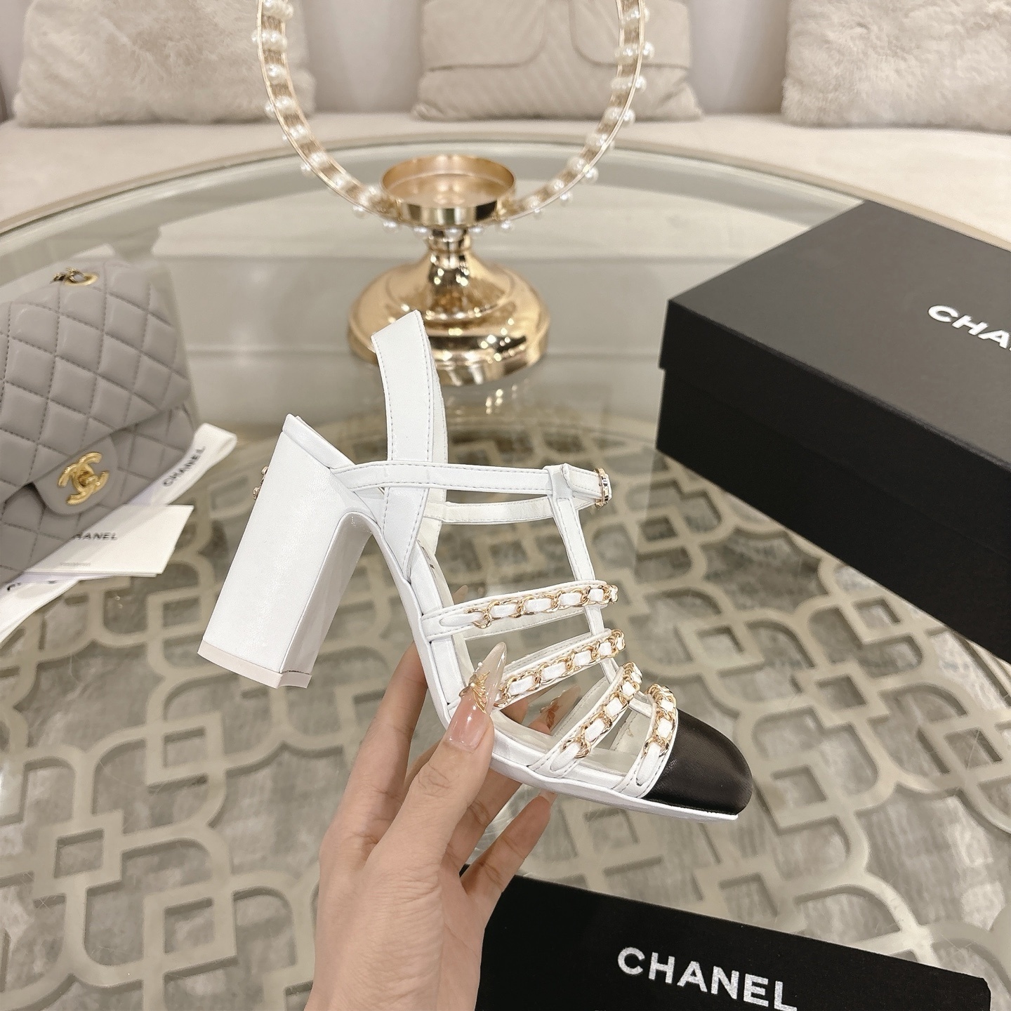 Elegant white leather sandals with black cap-toes and gold chain straps displayed on a glass table. - Hình 5