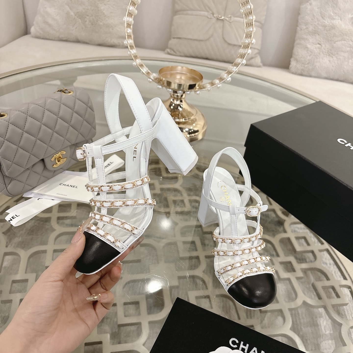 Elegant white leather sandals with black cap-toes and gold chain straps displayed on a glass table. - Hình 4