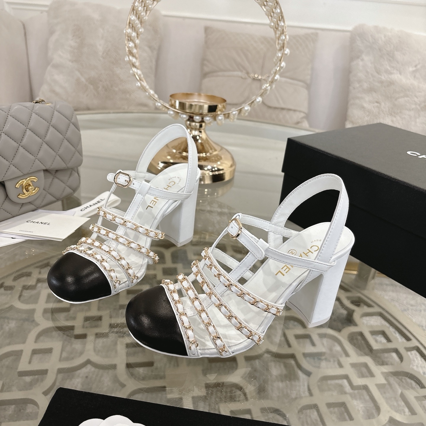 Elegant white leather sandals with black cap-toes and gold chain straps displayed on a glass table. - Hình 3
