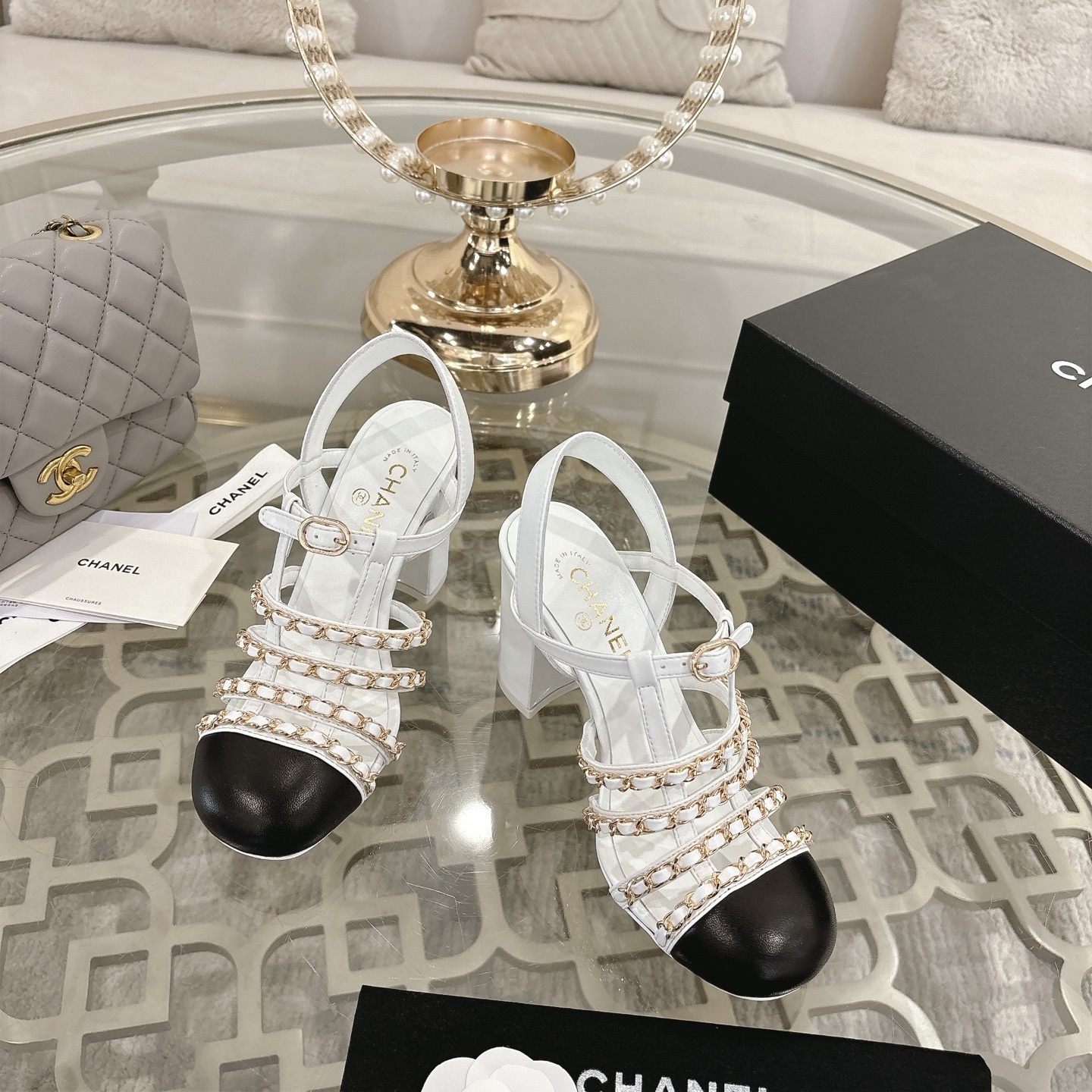 Elegant white leather sandals with black cap-toes and gold chain straps displayed on a glass table. - Hình 2
