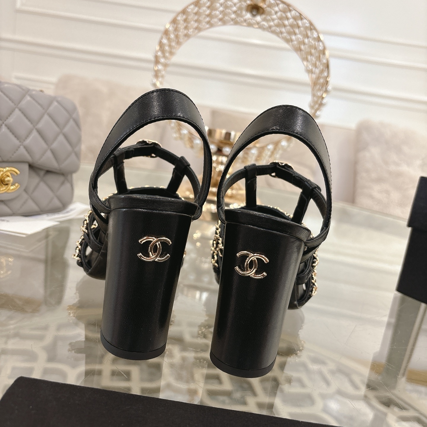 Pair of Chanel black leather sandals with gold chain straps and block heels displayed on a decorative glass table. - Hình 8
