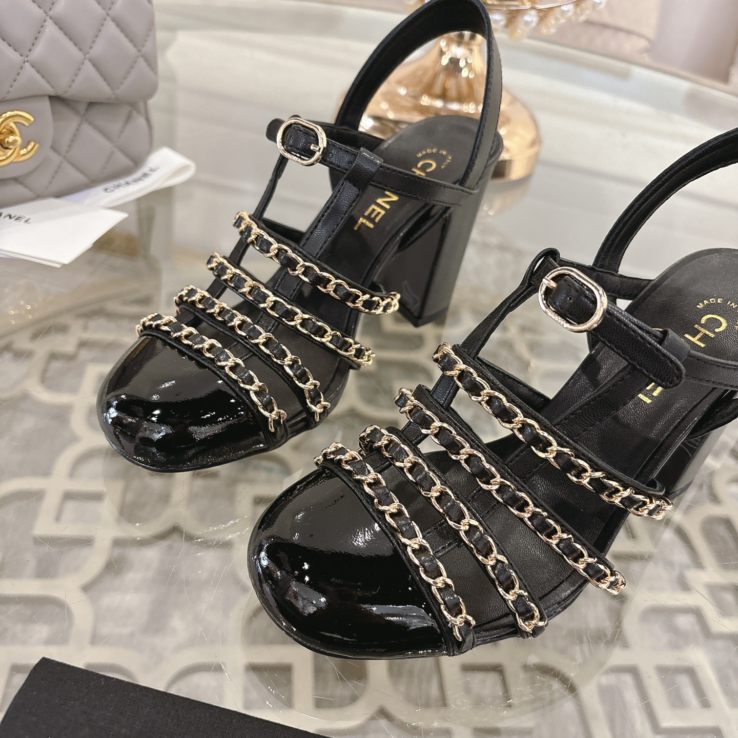 Pair of Chanel black leather sandals with gold chain straps and block heels displayed on a decorative glass table. - Hình 7