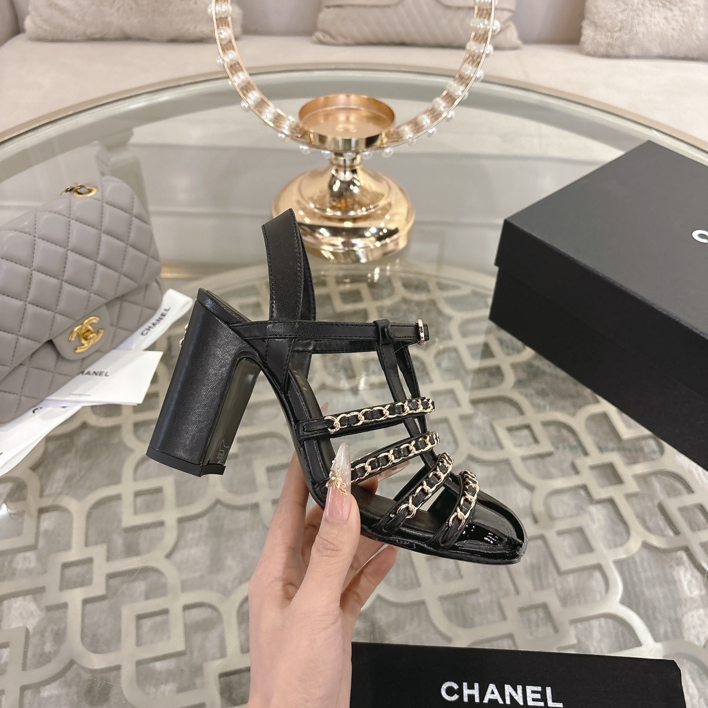 Pair of Chanel black leather sandals with gold chain straps and block heels displayed on a decorative glass table. - Hình 5