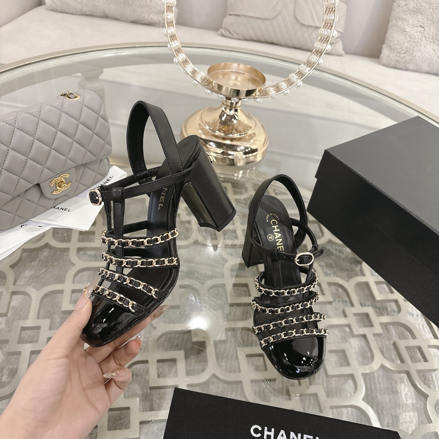 Pair of Chanel black leather sandals with gold chain straps and block heels displayed on a decorative glass table. - Hình 4