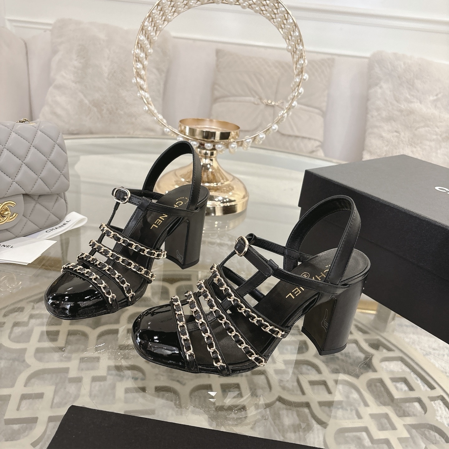 Pair of Chanel black leather sandals with gold chain straps and block heels displayed on a decorative glass table. - Hình 3
