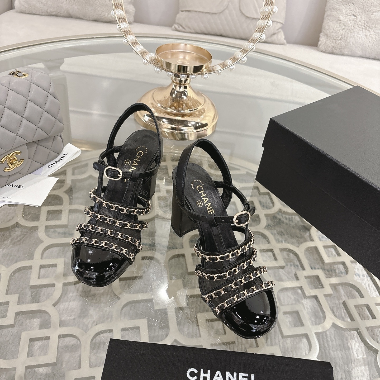 Pair of Chanel black leather sandals with gold chain straps and block heels displayed on a decorative glass table. - Hình 2