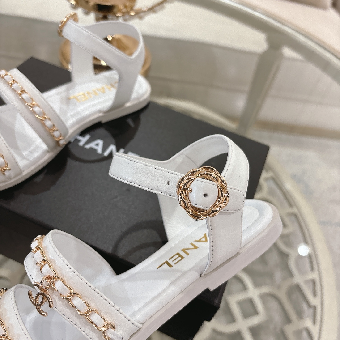 A pair of Chanel white leather flat sandals with gold chain link straps and CC logo charm displayed on a glass table. - Hình 8