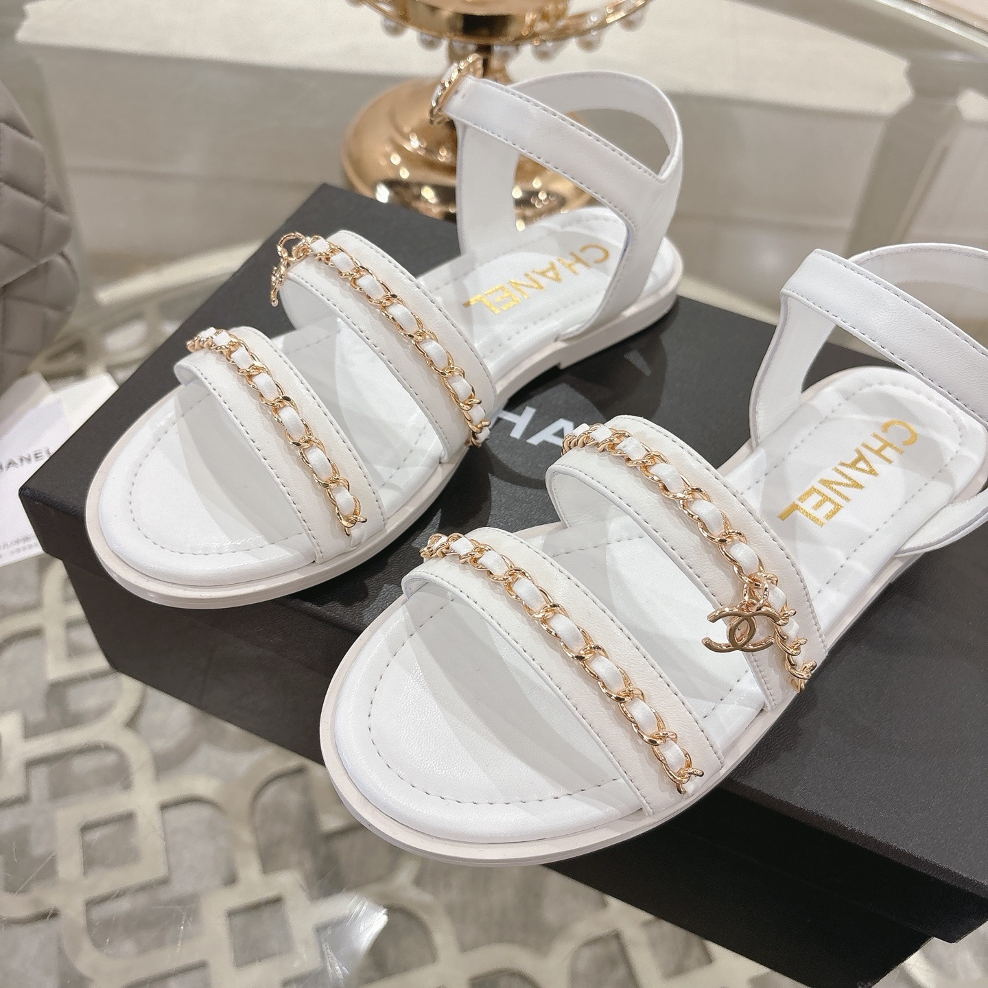 A pair of Chanel white leather flat sandals with gold chain link straps and CC logo charm displayed on a glass table. - Hình 7