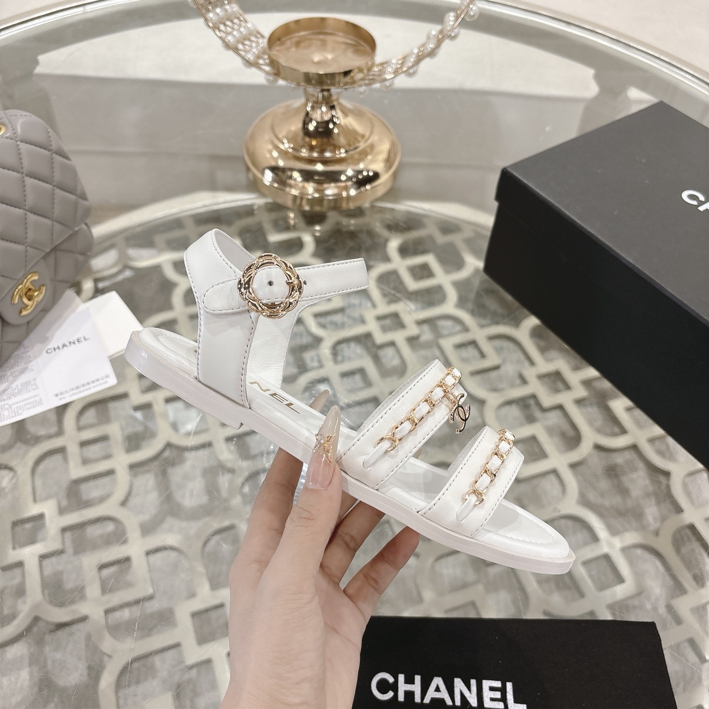 A pair of Chanel white leather flat sandals with gold chain link straps and CC logo charm displayed on a glass table. - Hình 5