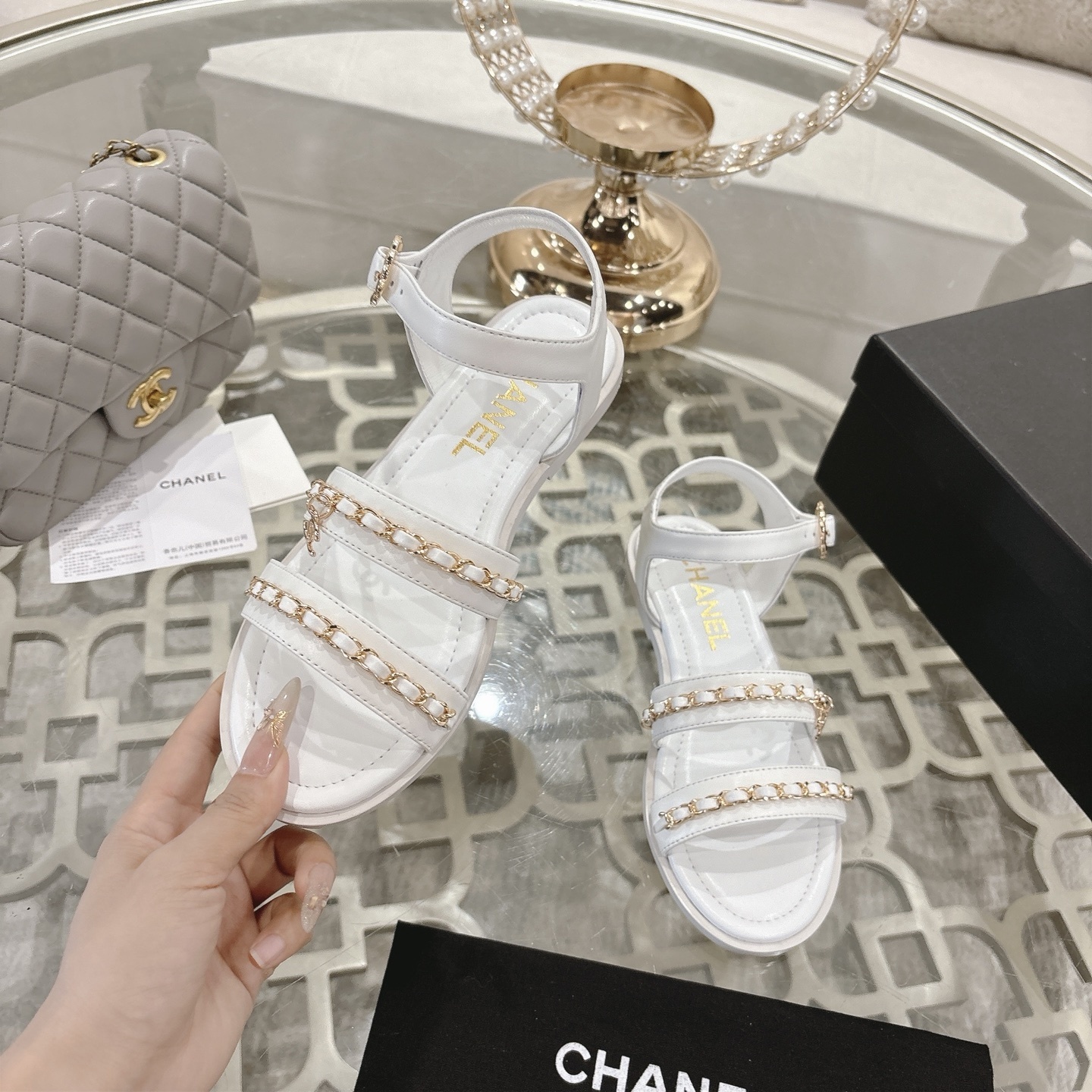 A pair of Chanel white leather flat sandals with gold chain link straps and CC logo charm displayed on a glass table. - Hình 4