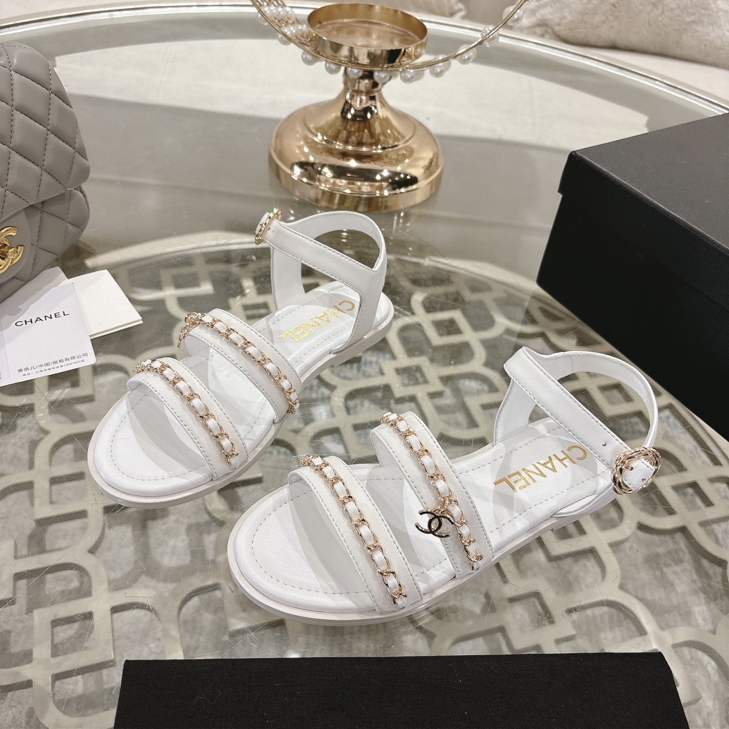 A pair of Chanel white leather flat sandals with gold chain link straps and CC logo charm displayed on a glass table. - Hình 3