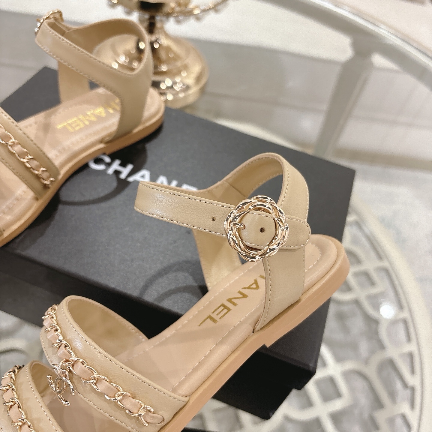 Pair of Chanel beige leather flat sandals with gold chain straps and ankle buckle displayed on a decorative glass table. - Hình 8