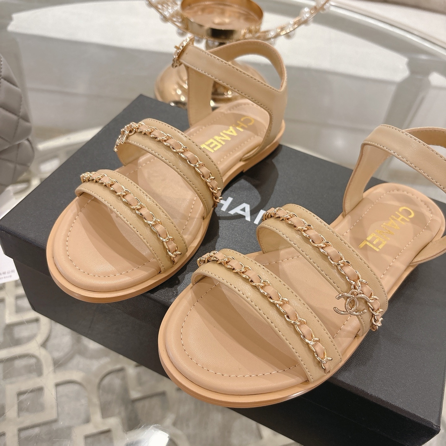 Pair of Chanel beige leather flat sandals with gold chain straps and ankle buckle displayed on a decorative glass table. - Hình 7