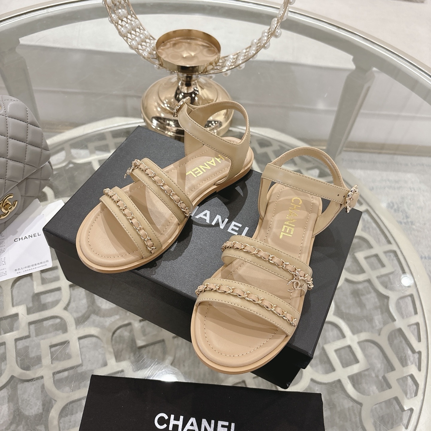 Pair of Chanel beige leather flat sandals with gold chain straps and ankle buckle displayed on a decorative glass table. - Hình 6