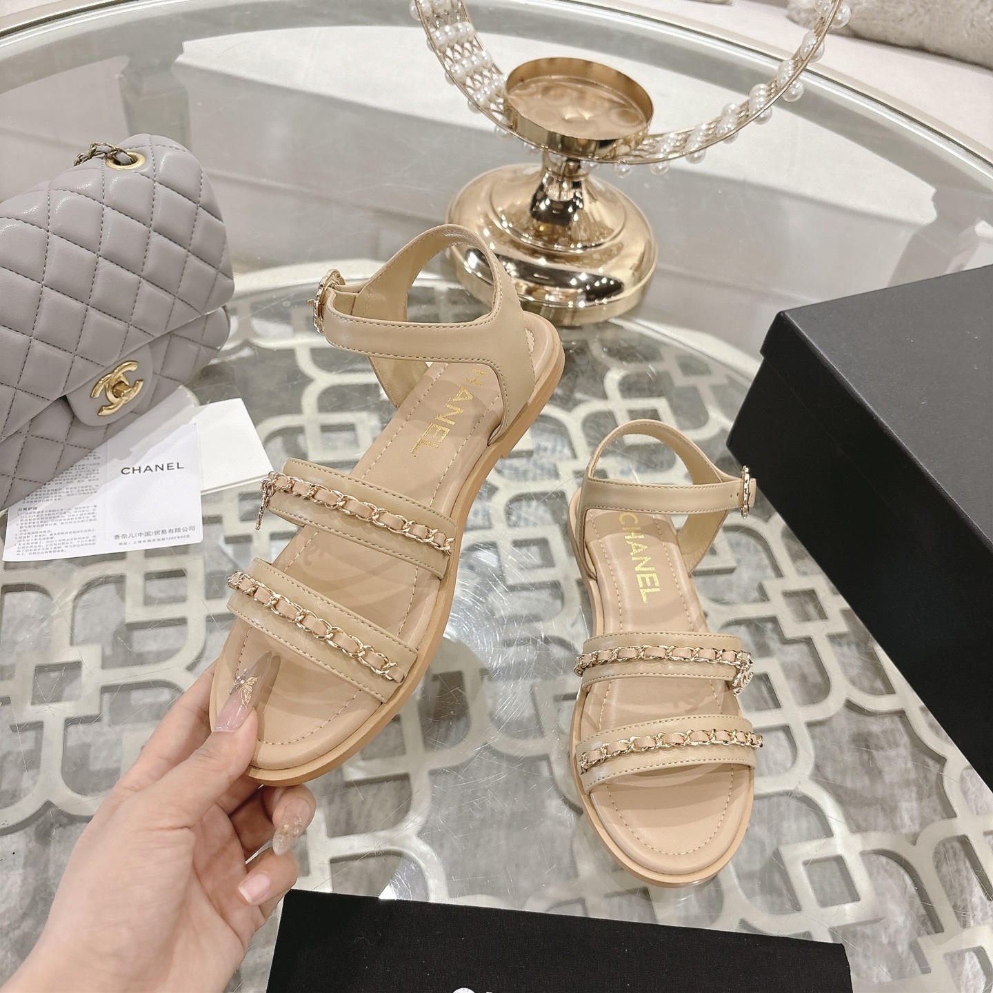Pair of Chanel beige leather flat sandals with gold chain straps and ankle buckle displayed on a decorative glass table. - Hình 4