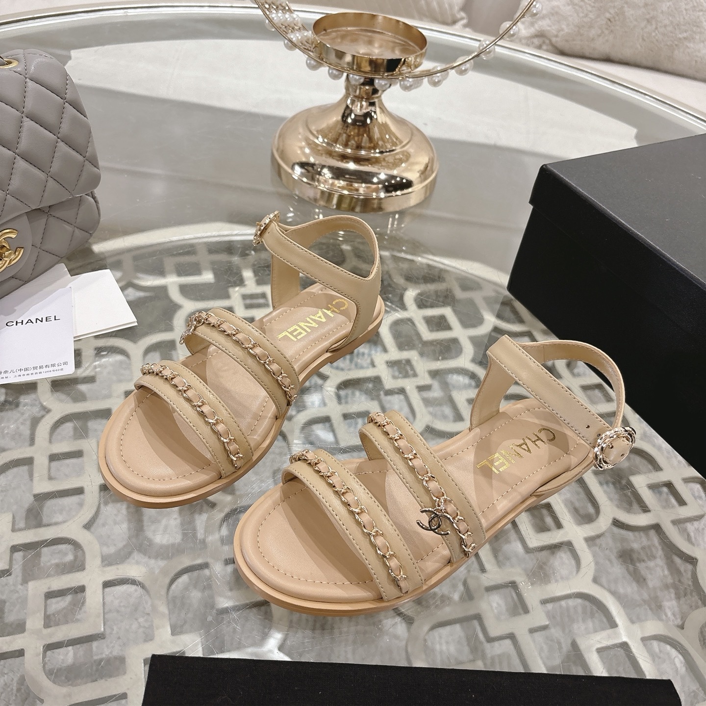 Pair of Chanel beige leather flat sandals with gold chain straps and ankle buckle displayed on a decorative glass table. - Hình 3