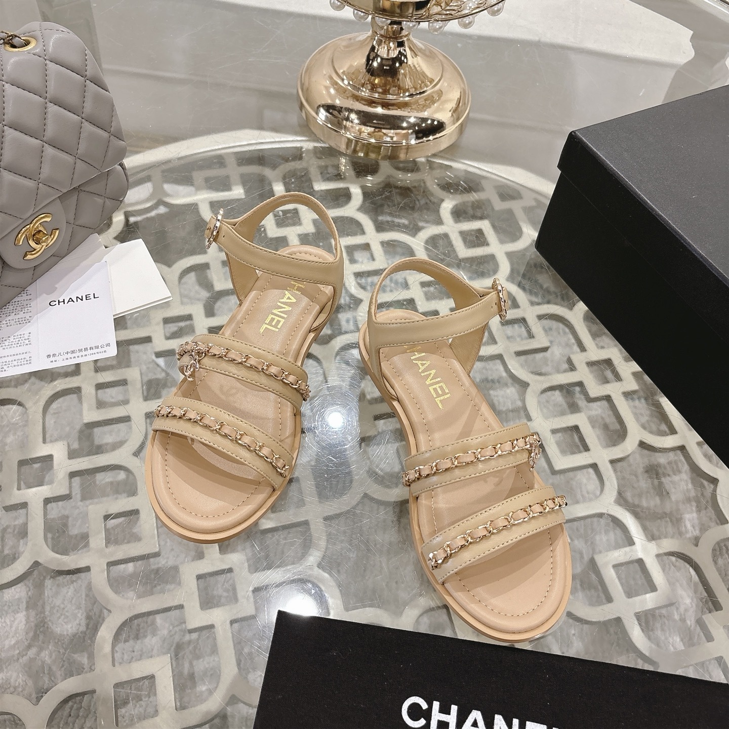 Pair of Chanel beige leather flat sandals with gold chain straps and ankle buckle displayed on a decorative glass table. - Hình 2