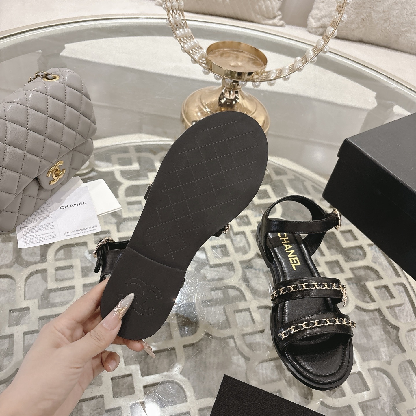 A pair of black Chanel flat sandals with gold chain straps and ankle buckles on a glass table - Hình 9