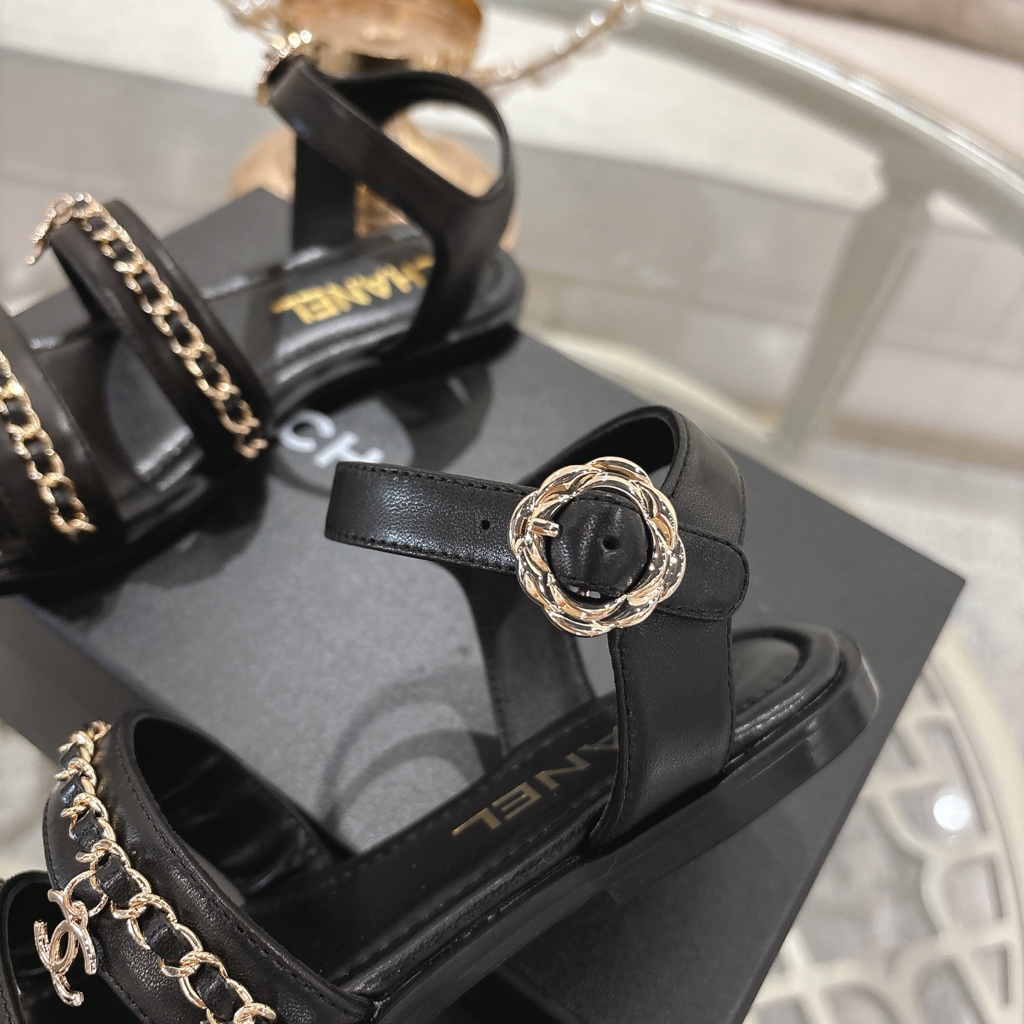 A pair of black Chanel flat sandals with gold chain straps and ankle buckles on a glass table - Hình 8