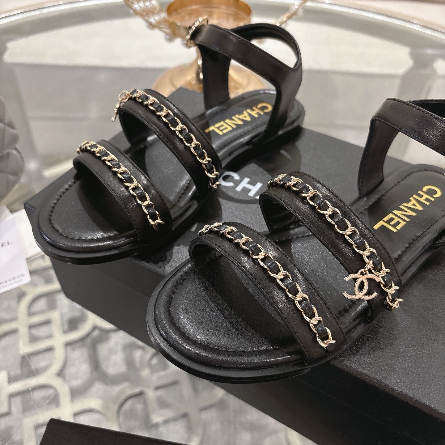 A pair of black Chanel flat sandals with gold chain straps and ankle buckles on a glass table - Hình 7