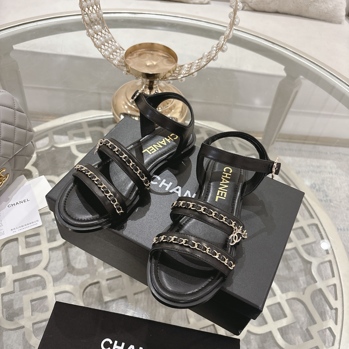 A pair of black Chanel flat sandals with gold chain straps and ankle buckles on a glass table - Hình 6