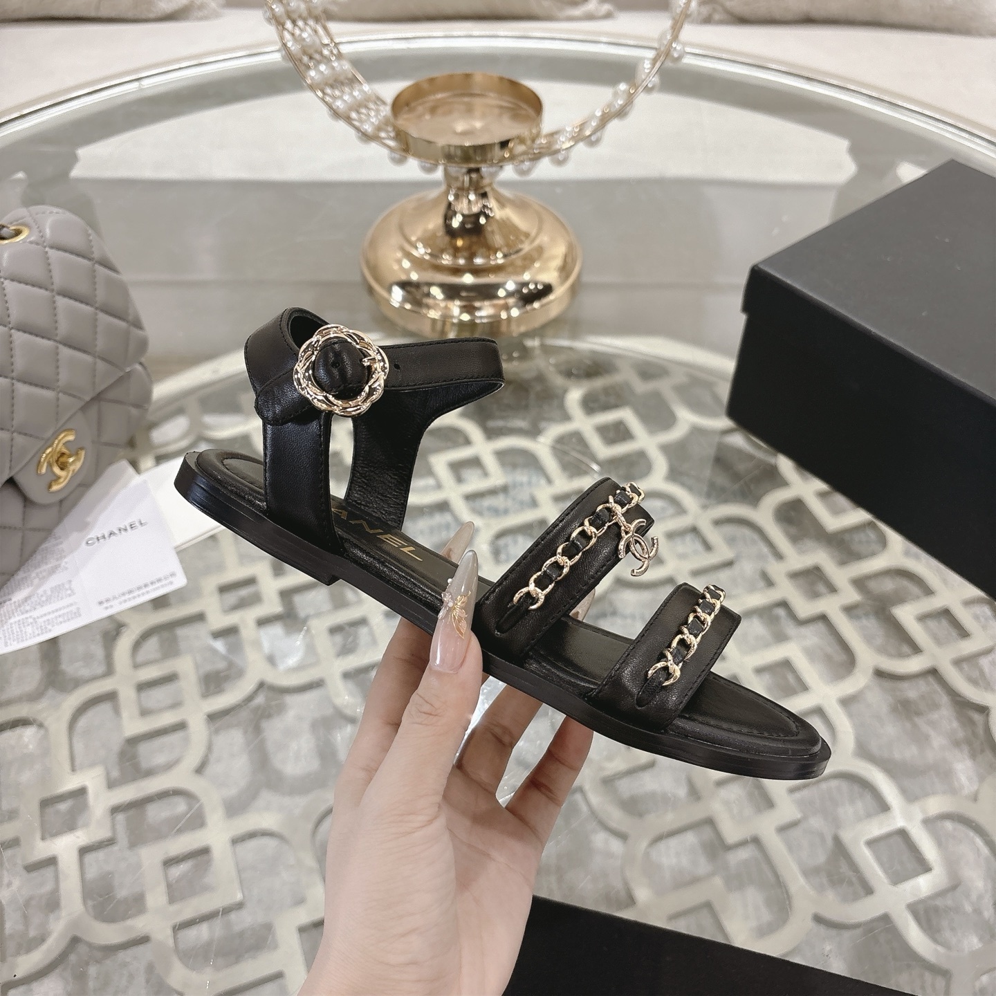 A pair of black Chanel flat sandals with gold chain straps and ankle buckles on a glass table - Hình 5