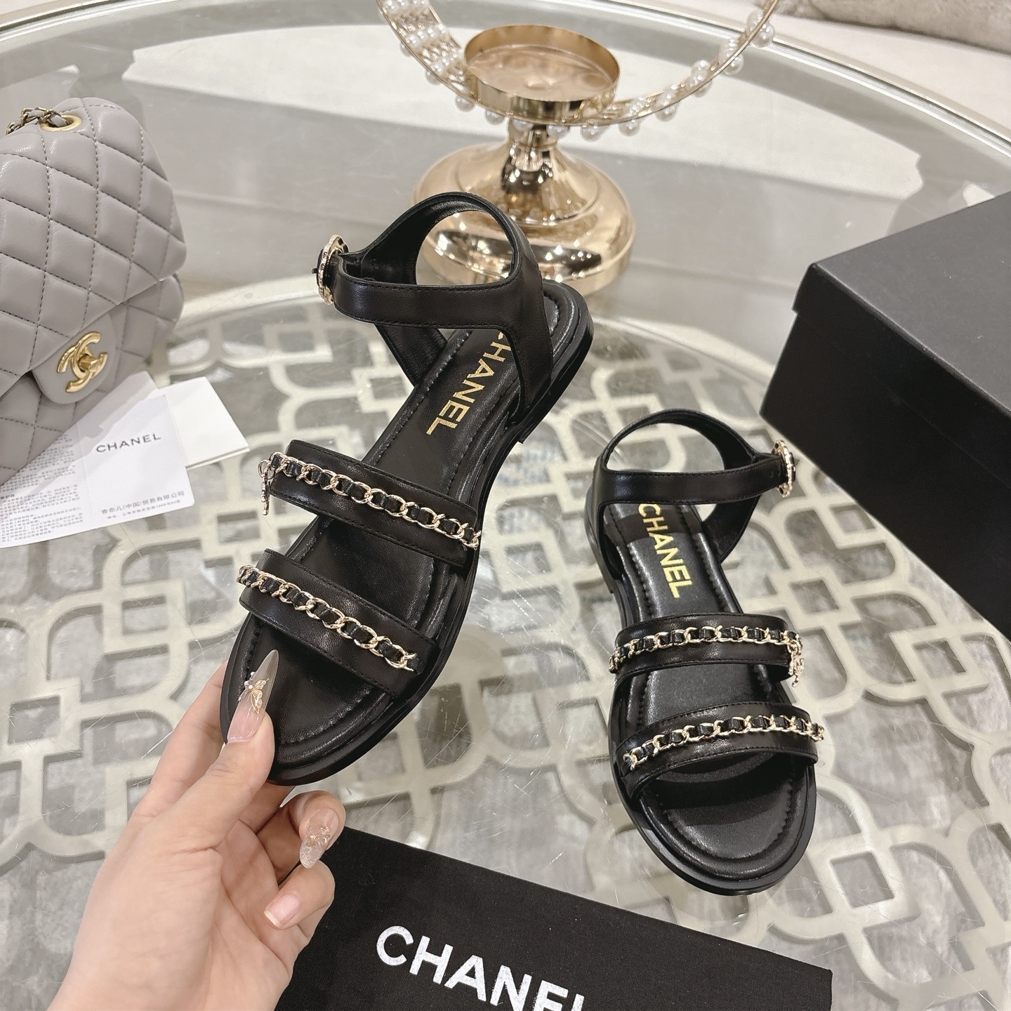 A pair of black Chanel flat sandals with gold chain straps and ankle buckles on a glass table - Hình 4