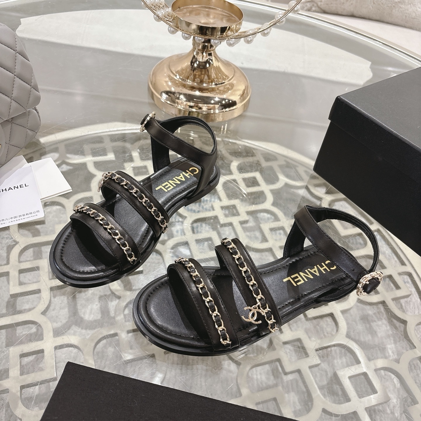 A pair of black Chanel flat sandals with gold chain straps and ankle buckles on a glass table - Hình 3