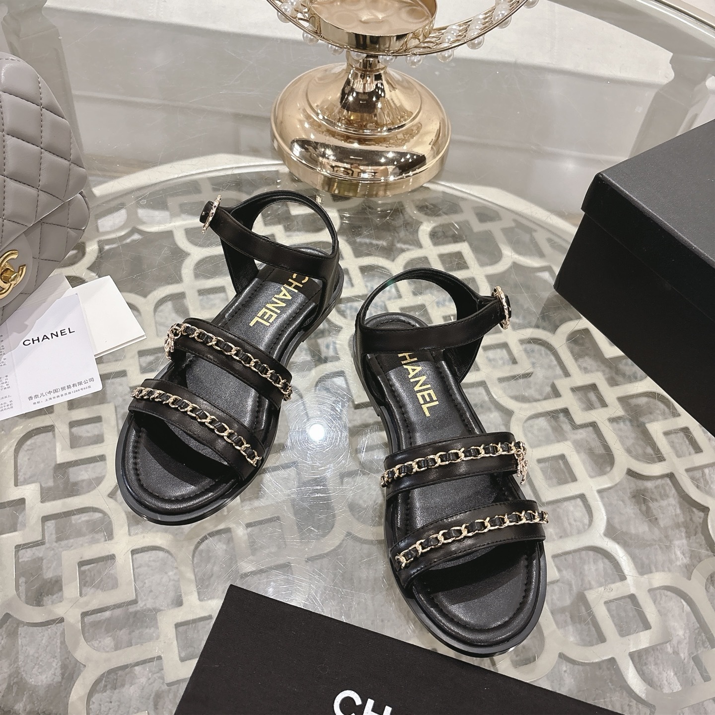 A pair of black Chanel flat sandals with gold chain straps and ankle buckles on a glass table - Hình 2