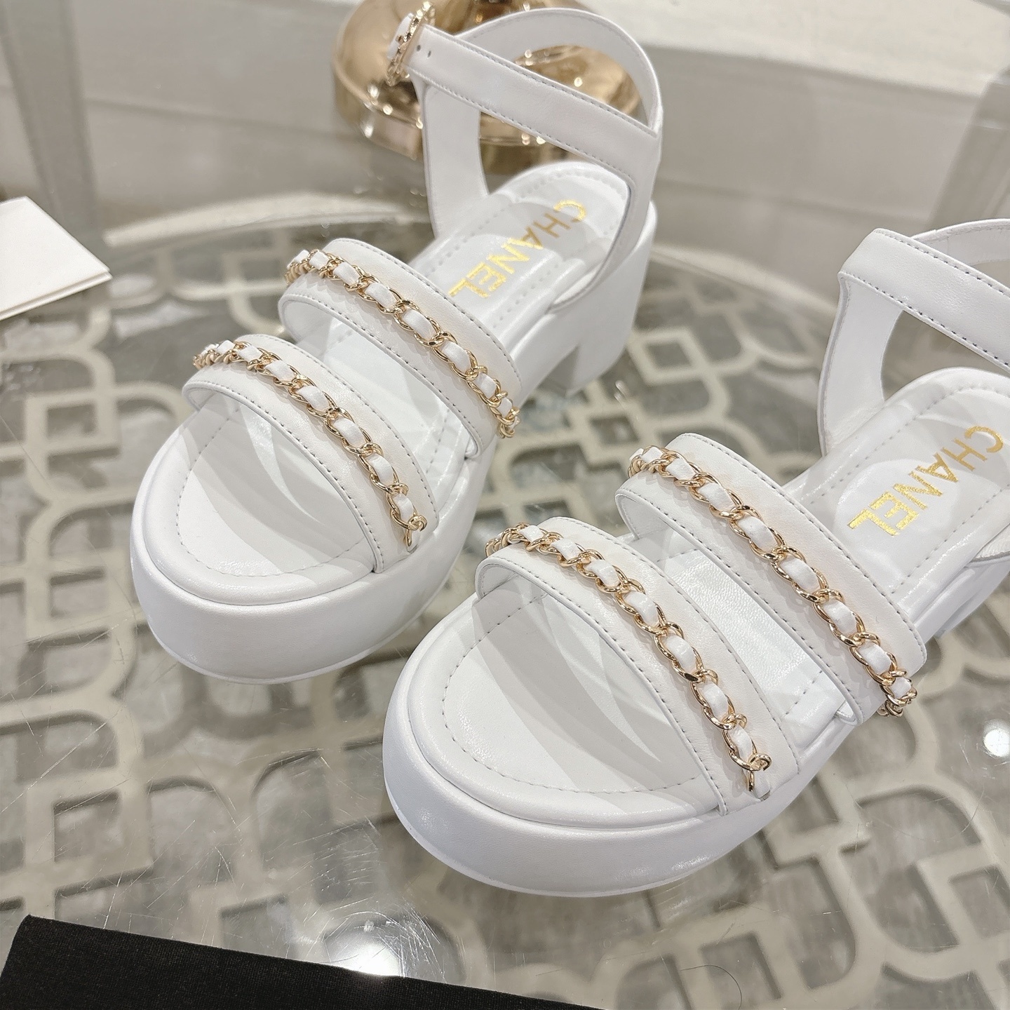 Elegant white platform sandals with gold chain-link straps and adjustable ankle buckles displayed on a glass table. - Hình 7