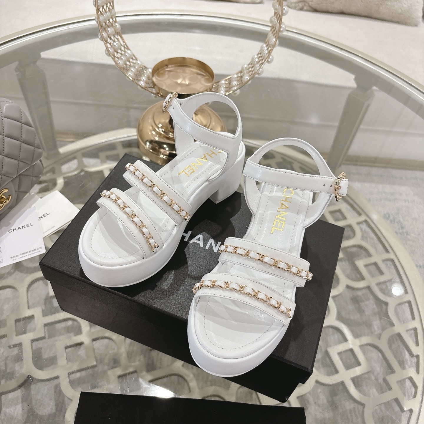 Elegant white platform sandals with gold chain-link straps and adjustable ankle buckles displayed on a glass table. - Hình 6