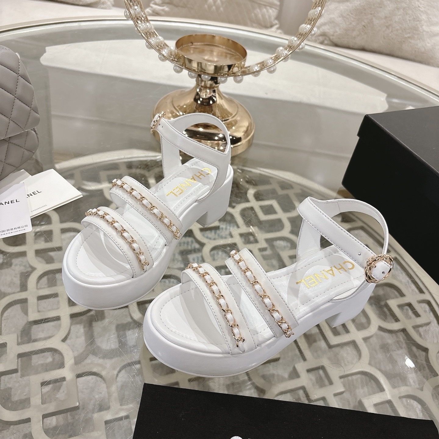 Elegant white platform sandals with gold chain-link straps and adjustable ankle buckles displayed on a glass table. - Hình 3