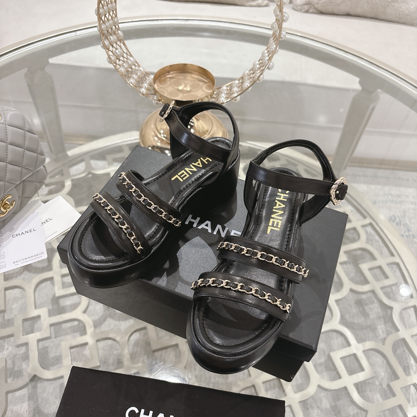 Elegant black platform sandals with two gold chain straps and an ankle buckle displayed on a decorative glass table. - Hình 6