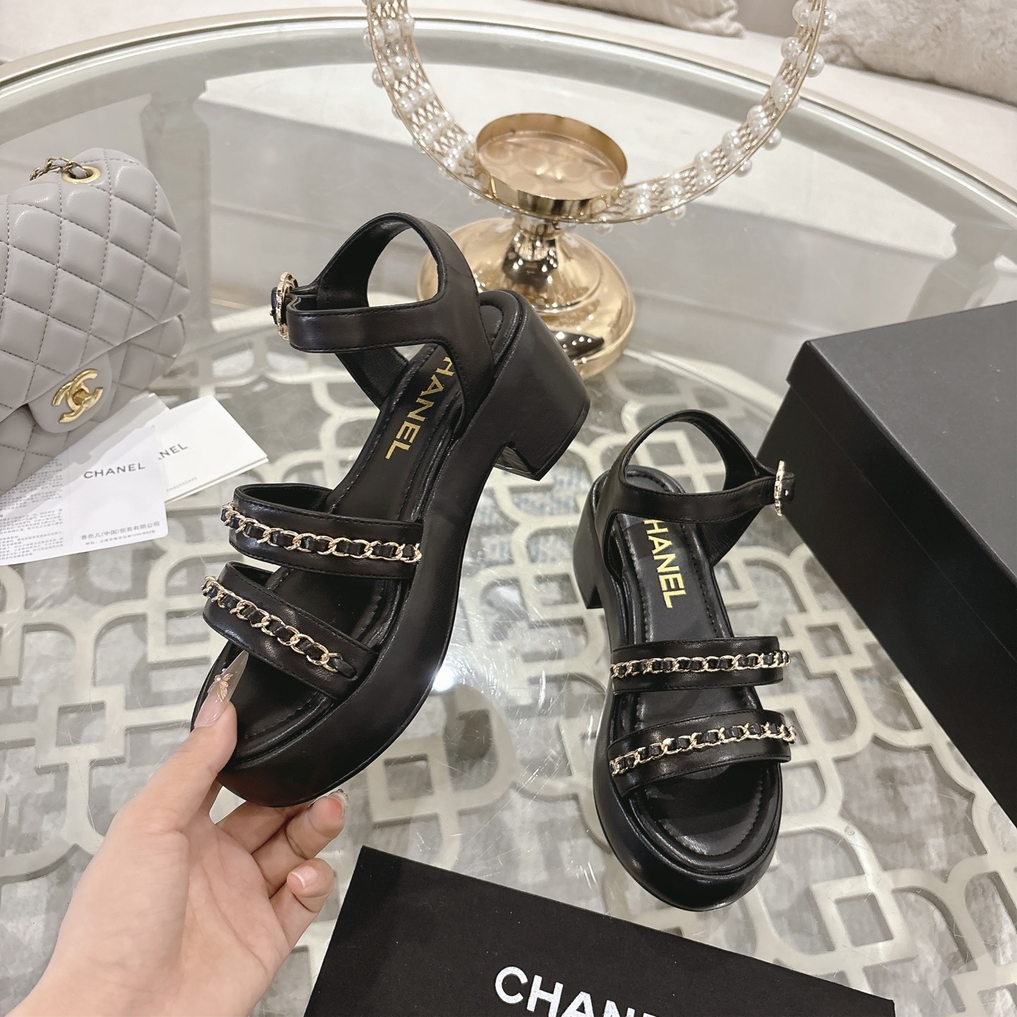 Elegant black platform sandals with two gold chain straps and an ankle buckle displayed on a decorative glass table. - Hình 4