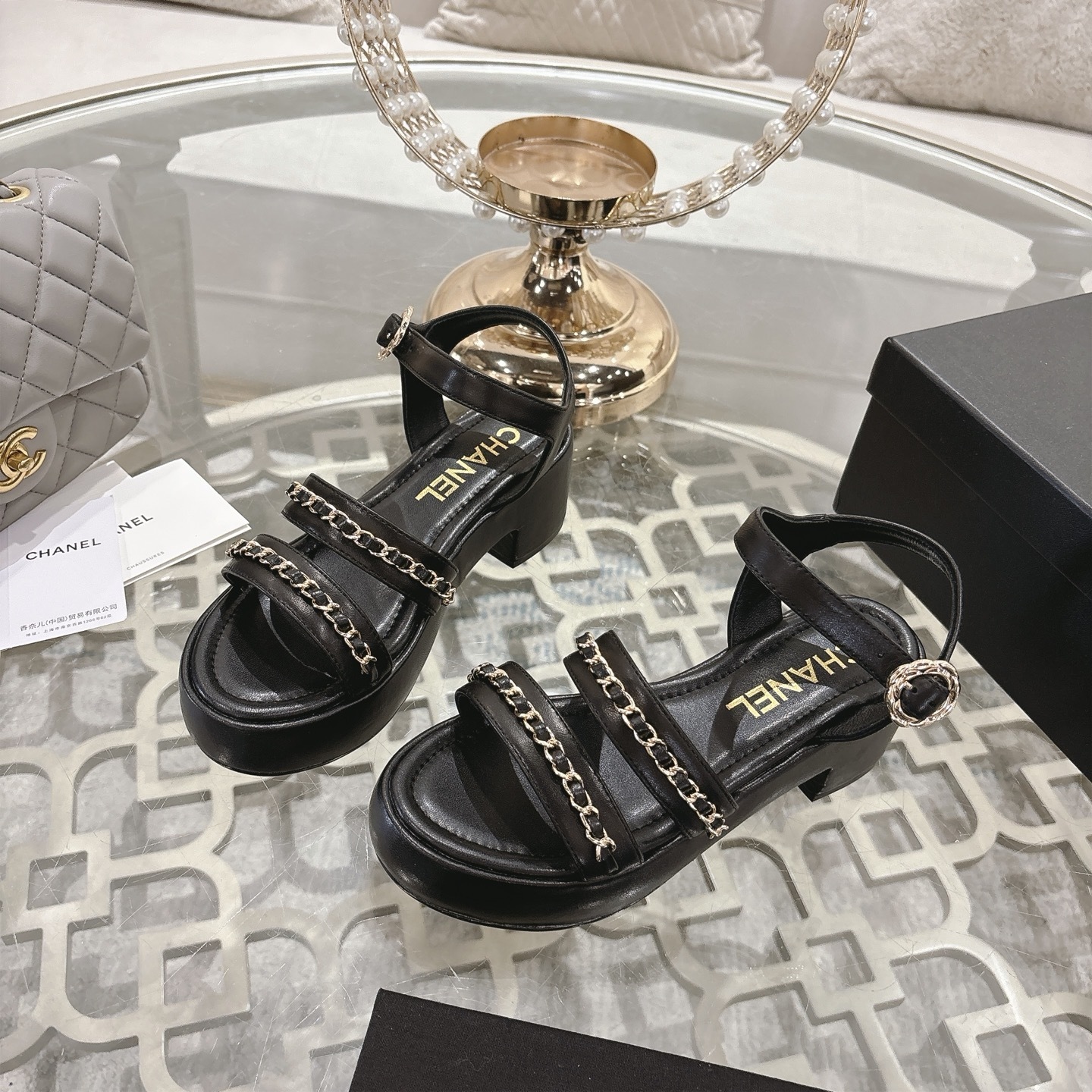 Elegant black platform sandals with two gold chain straps and an ankle buckle displayed on a decorative glass table. - Hình 3