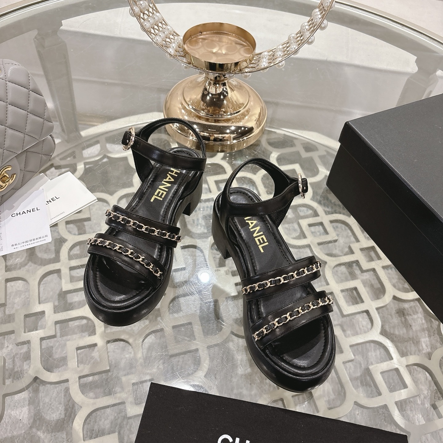 Elegant black platform sandals with two gold chain straps and an ankle buckle displayed on a decorative glass table. - Hình 2