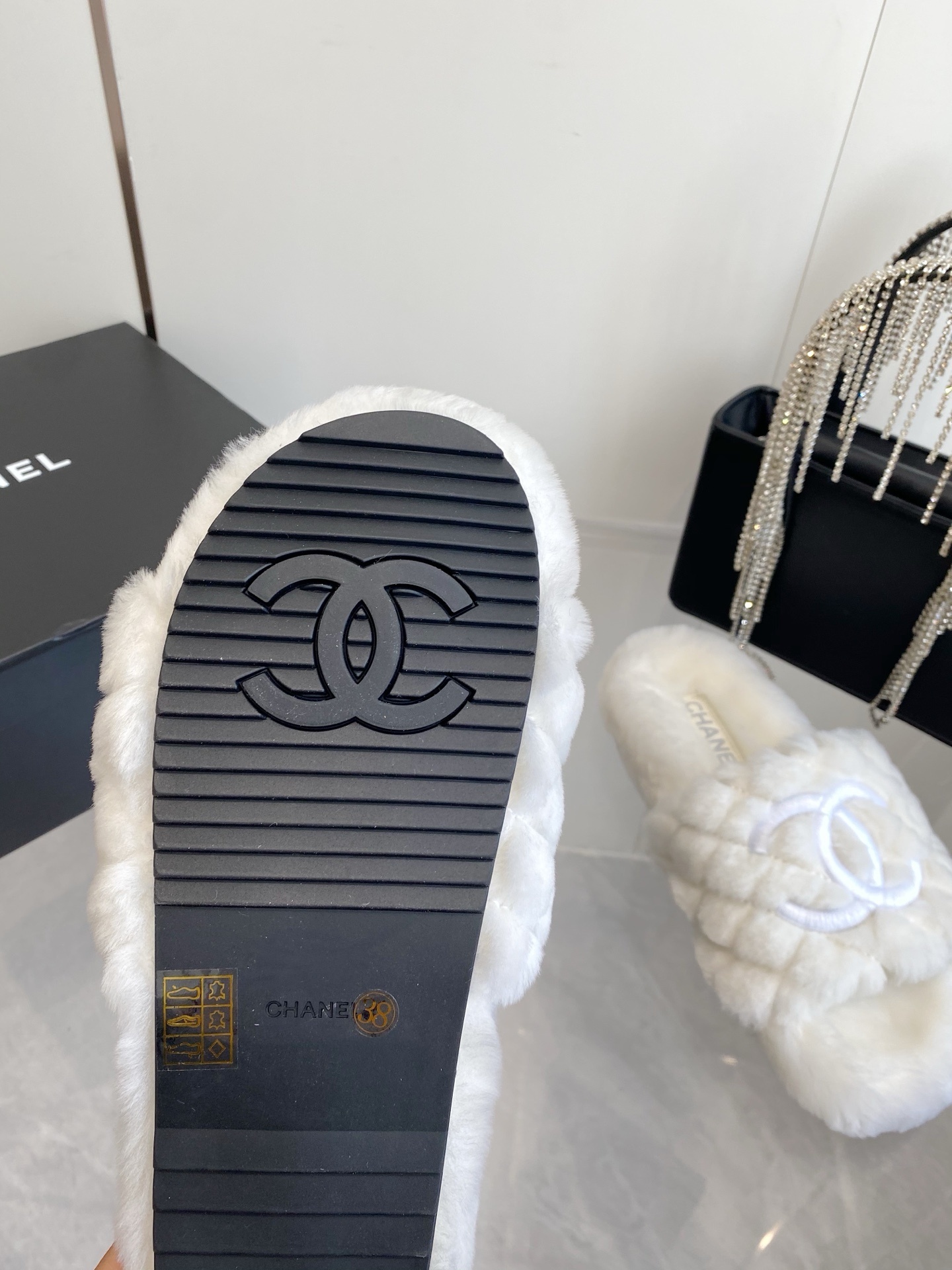 A pair of luxury white quilted faux fur slippers with an embroidered logo on a marble surface next to a black designer bag. - Hình 9