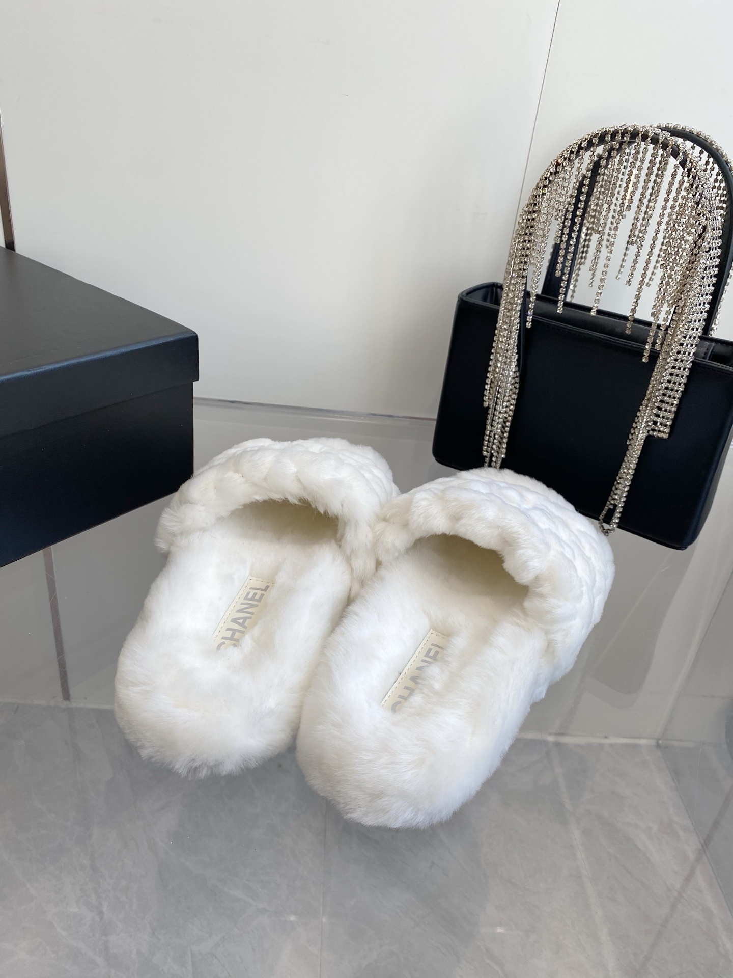 A pair of luxury white quilted faux fur slippers with an embroidered logo on a marble surface next to a black designer bag. - Hình 8