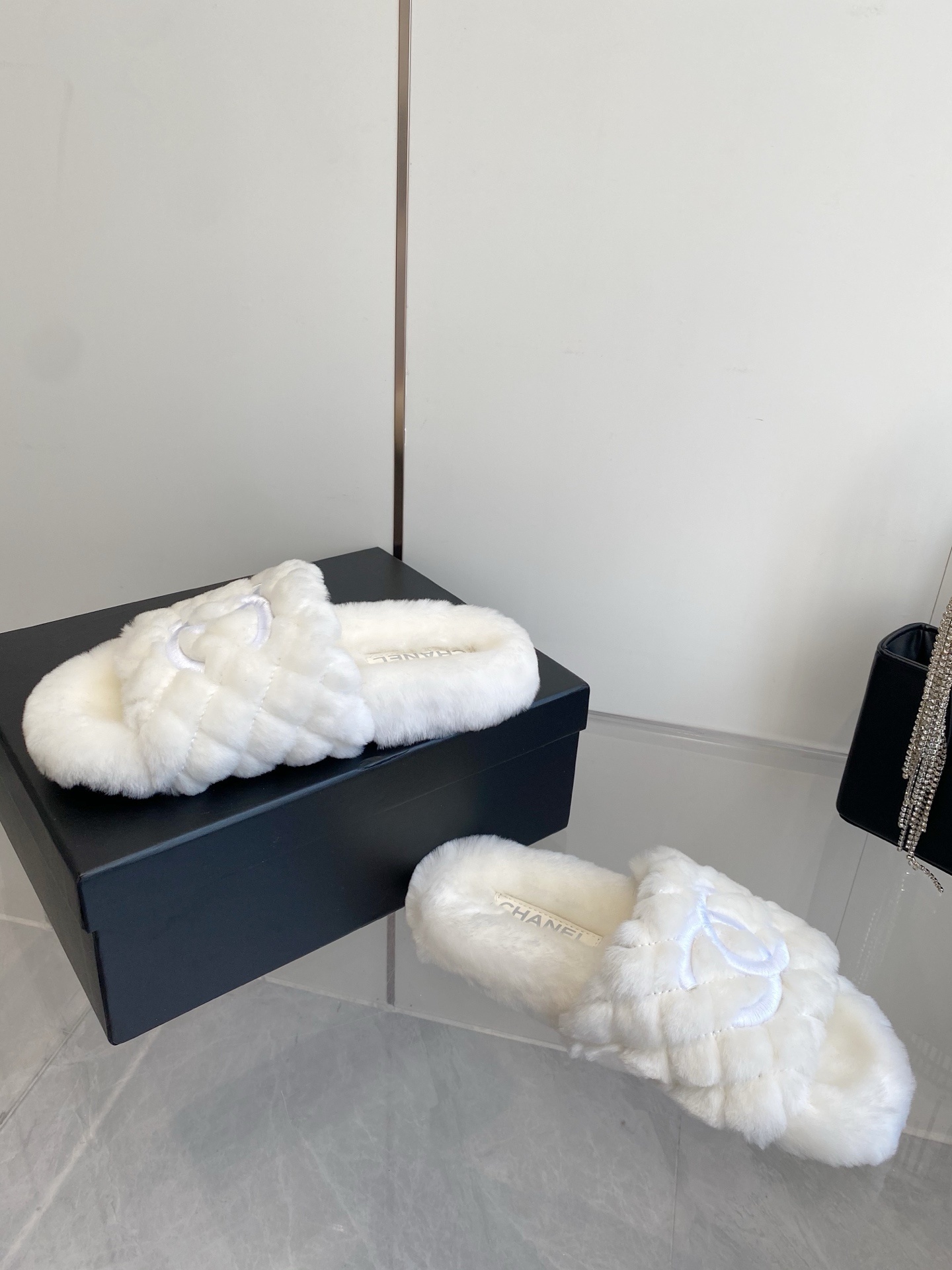 A pair of luxury white quilted faux fur slippers with an embroidered logo on a marble surface next to a black designer bag. - Hình 7