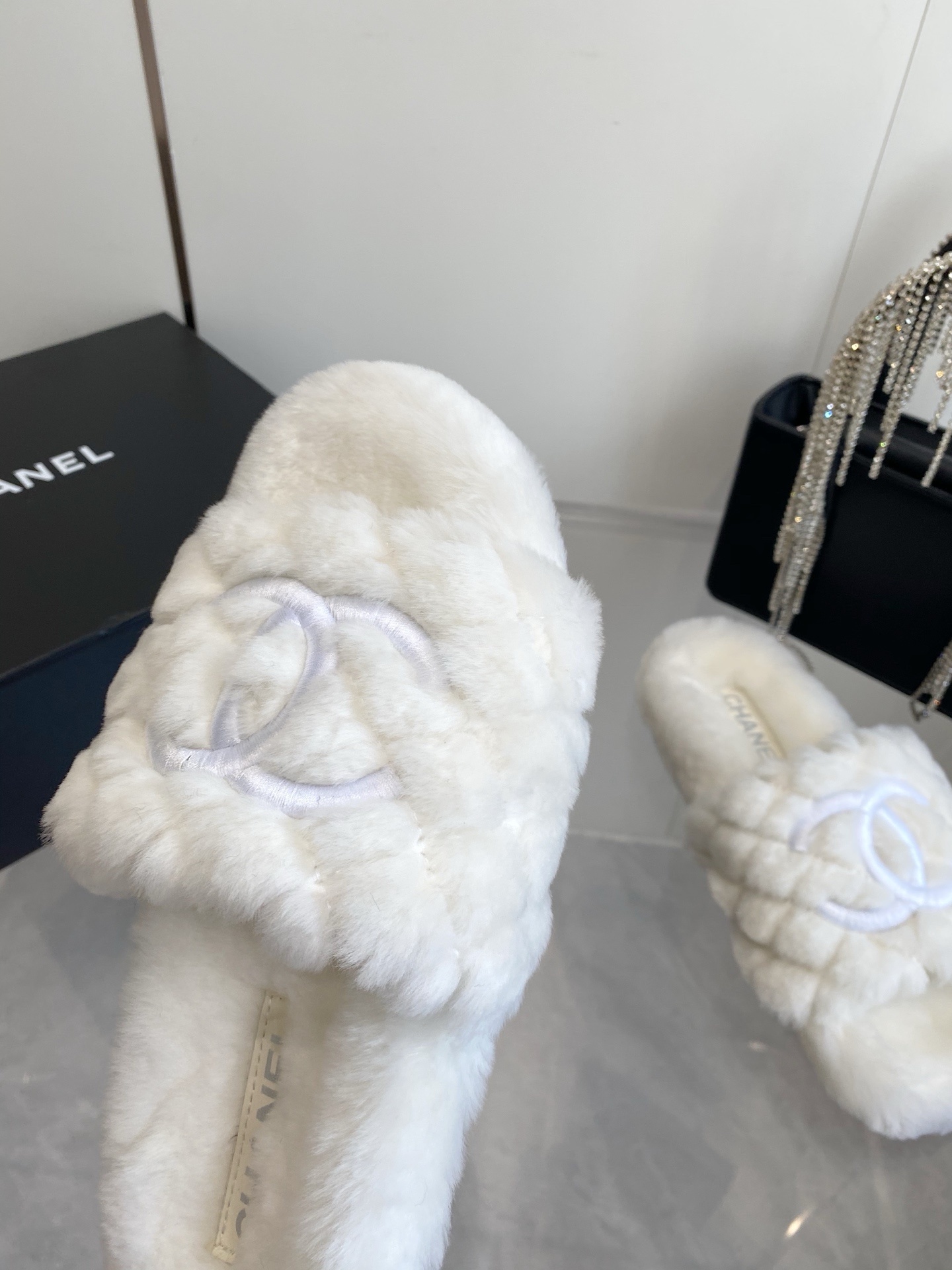 A pair of luxury white quilted faux fur slippers with an embroidered logo on a marble surface next to a black designer bag. - Hình 6