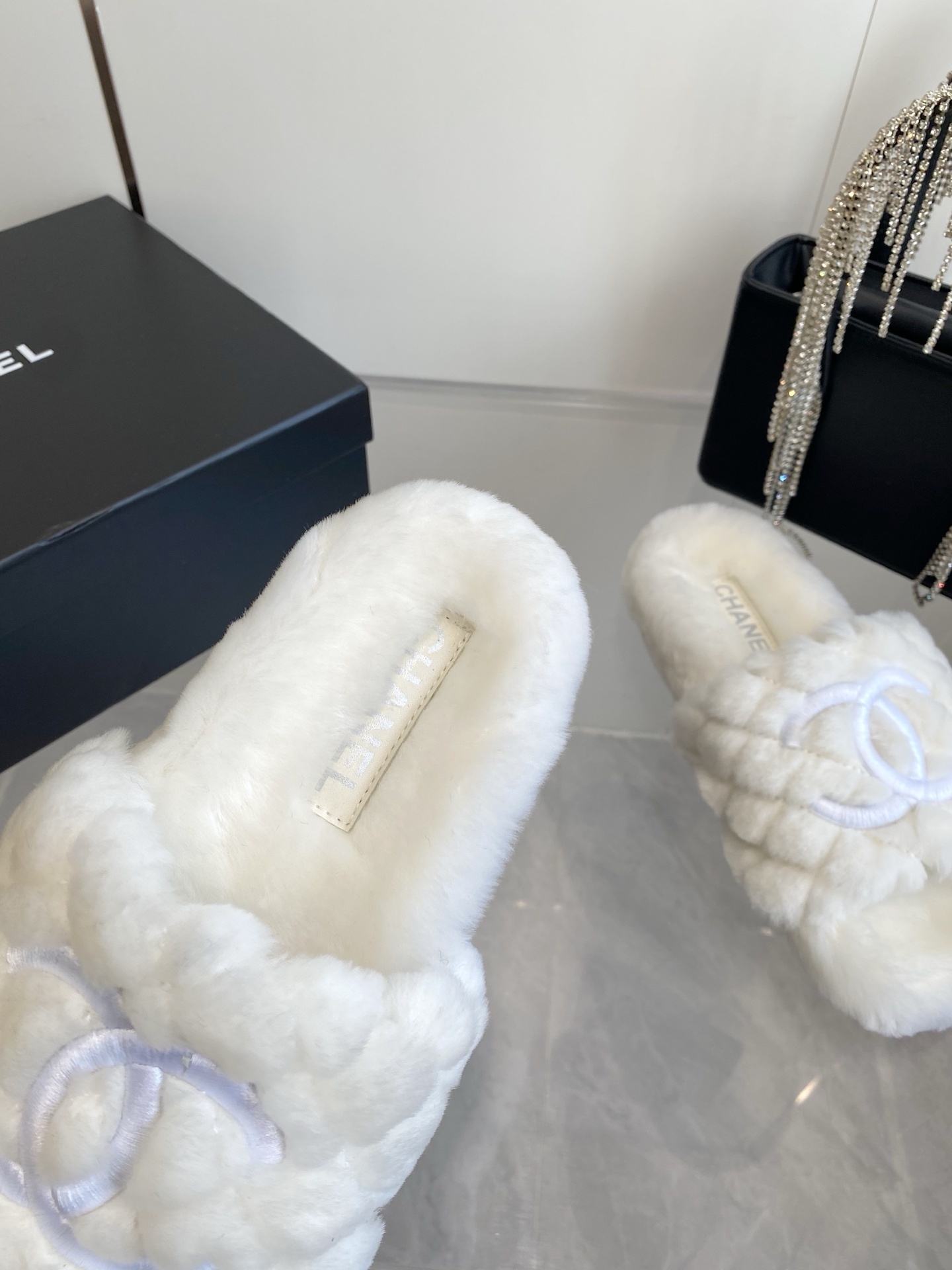 A pair of luxury white quilted faux fur slippers with an embroidered logo on a marble surface next to a black designer bag. - Hình 5