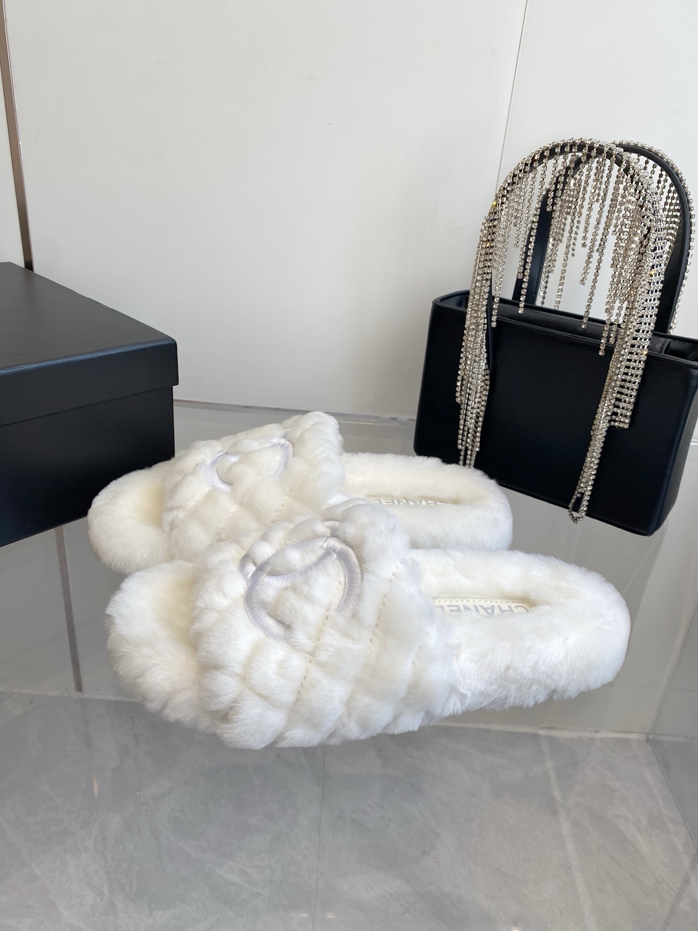 A pair of luxury white quilted faux fur slippers with an embroidered logo on a marble surface next to a black designer bag. - Hình 2