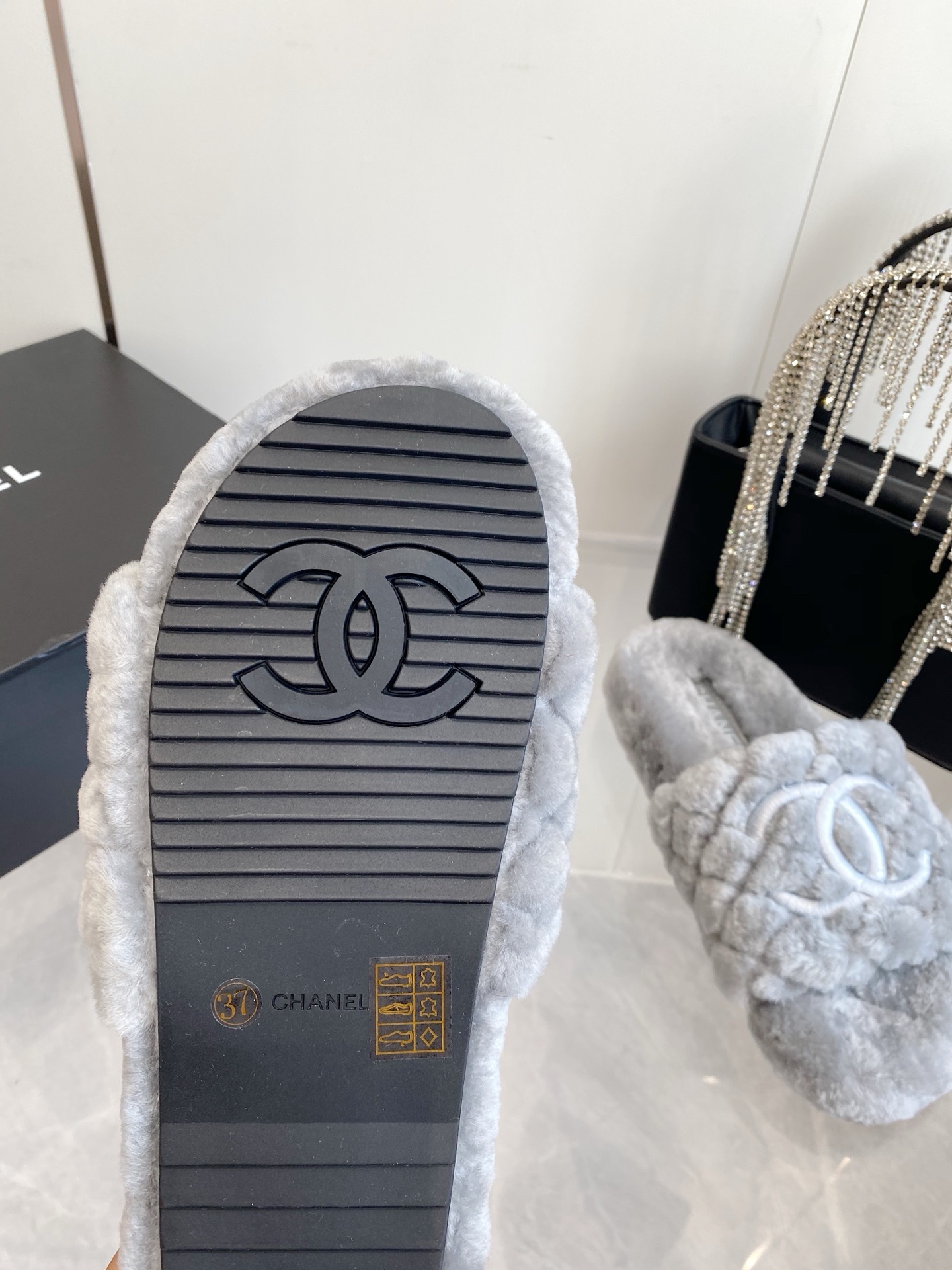 A pair of premium grey quilted faux fur slides featuring an interlocking logo design on a marble surface. - Hình 9