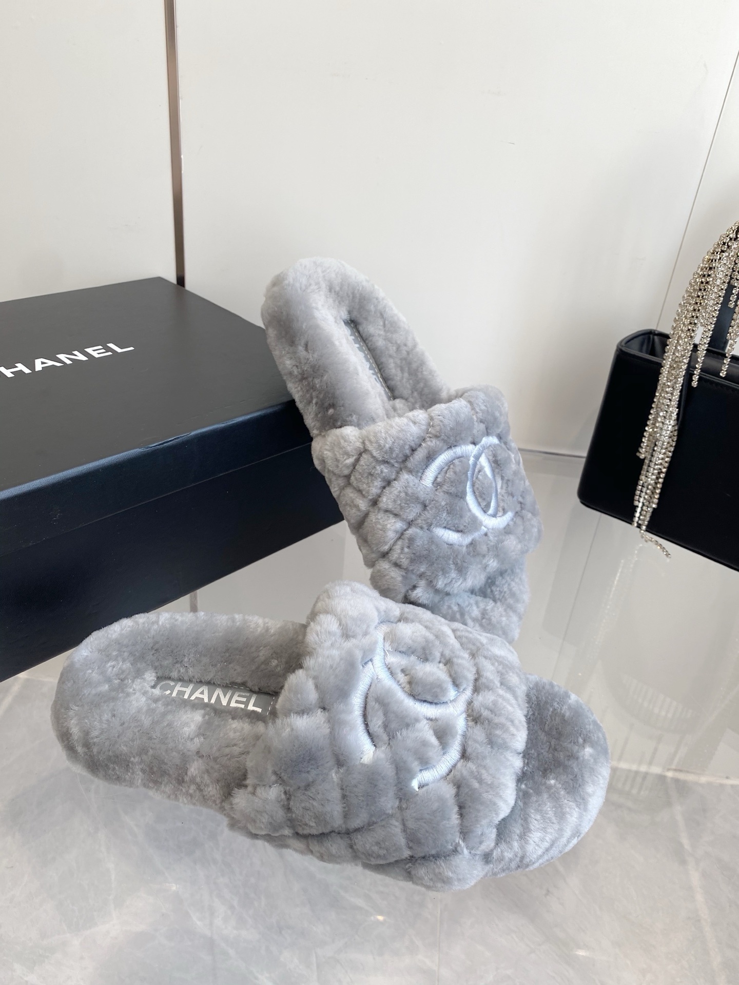 A pair of premium grey quilted faux fur slides featuring an interlocking logo design on a marble surface. - Hình 6