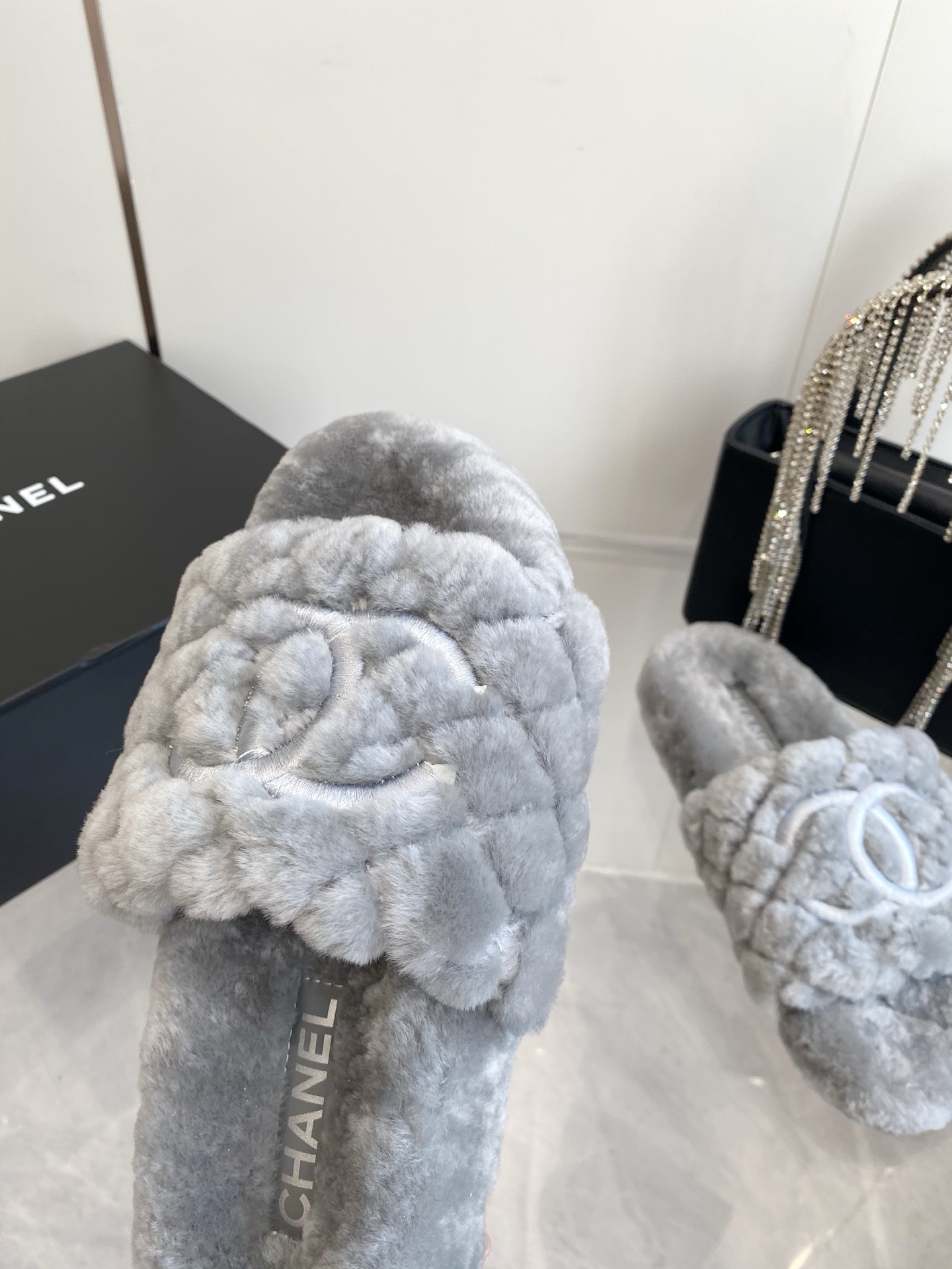 A pair of premium grey quilted faux fur slides featuring an interlocking logo design on a marble surface. - Hình 5